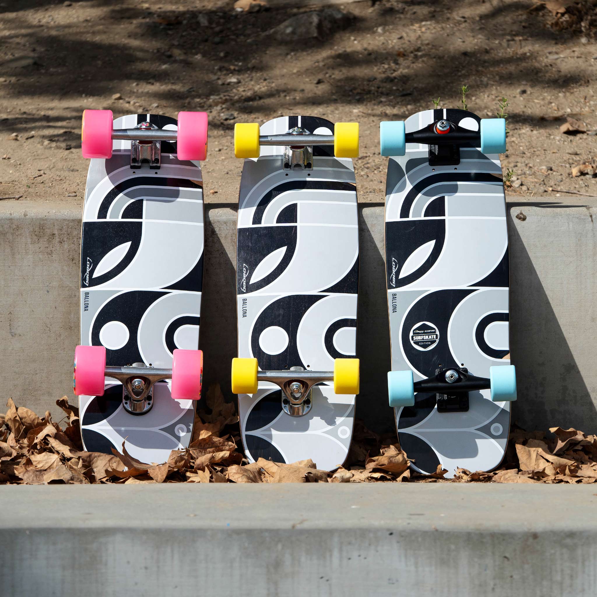 Three Loaded Ballona Orca longboards with a matching black, white, and gray abstract wave pattern are lined up on a concrete ledge outdoors. The boards feature different colored wheels: pink, yellow, and light blue.