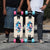 Two longboards are shown outdoors on a paved road, held upright by two partially visible people. The boards have a cream-colored deck with a colorful geometric graphic in the center, black trucks, and wheels in pink and turquoise.