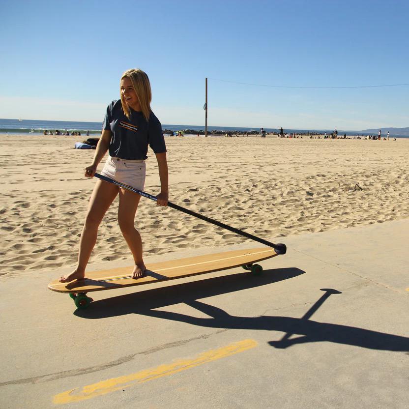A young woman riding a longboard along a sandy beach pathway.