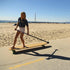 A young woman riding a longboard along a sandy beach pathway.