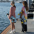 Two women standing on a dock by the water holding longboards. One wears a pink ribbed knit sweater dress and a tan beanie, and the other wears a pastel tie-dye t-shirt, black pants, and white sneakers.