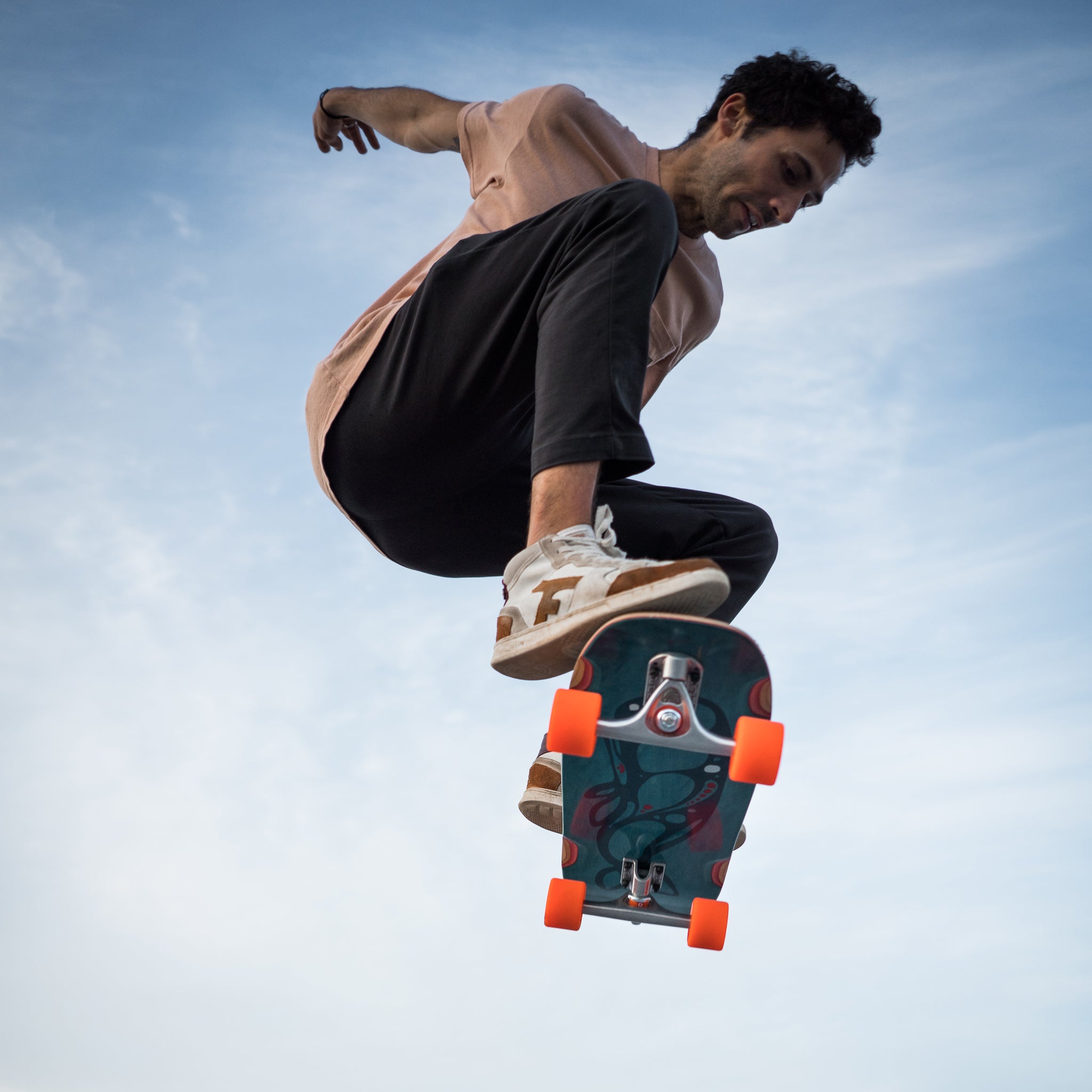 A skateboarder mid-air performing a trick against a light blue sky, wearing a light pink t-shirt, black pants, and white sneakers with orange wheels on the skateboard.