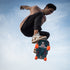 A skateboarder mid-air performing a trick against a light blue sky, wearing a light pink t-shirt, black pants, and white sneakers with orange wheels on the skateboard.