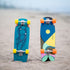 Two colorful longboards standing upright in sand on a beach, one with yellow wheels and a blue graphic design, and the other with orange wheels and a yellow, blue, and pink geometric design.