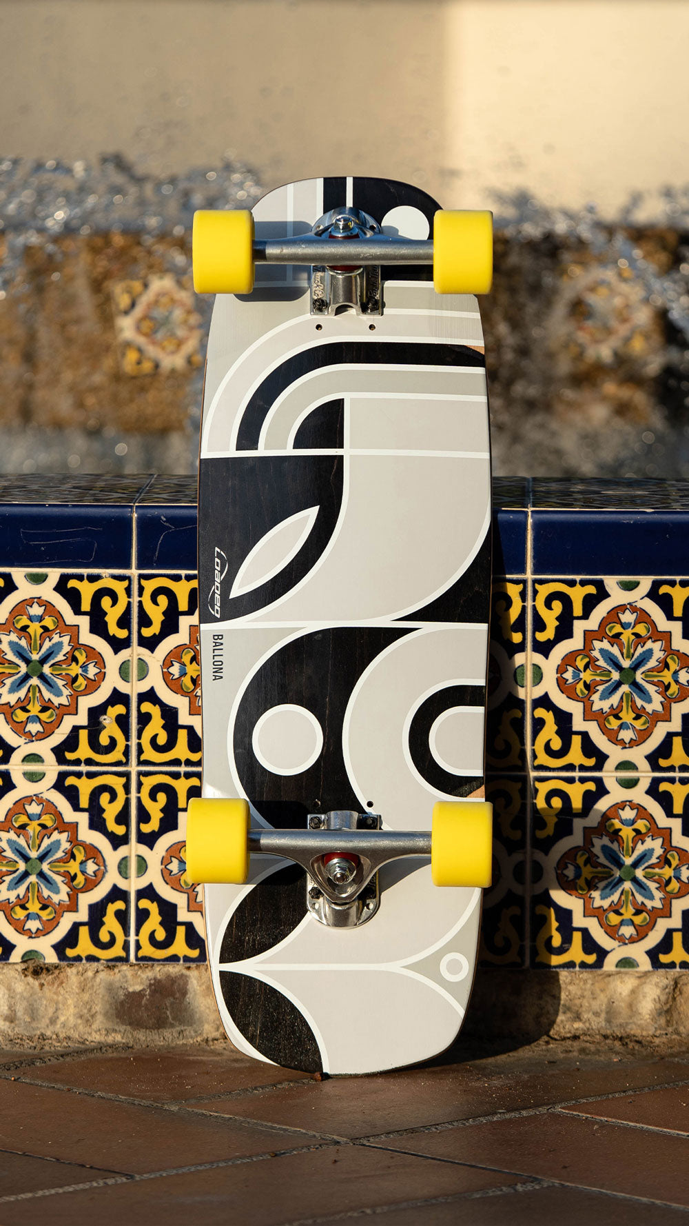 A longboard skateboard with a bold black and white graphic design and bright yellow wheels, leaning against a decorative tiled wall.