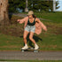 A woman skateboarding outdoors on a paved path next to a grassy park area, wearing a beige bucket hat, a dark green tank top, a light pink open cardigan, denim shorts, and white sneakers.