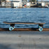 A Loaded Ballona Orca Surfskate Edition longboard skateboard with a black grip tape deck, black trucks, and light blue wheels resting on a wooden dock with a blurred harbor and water in the background.