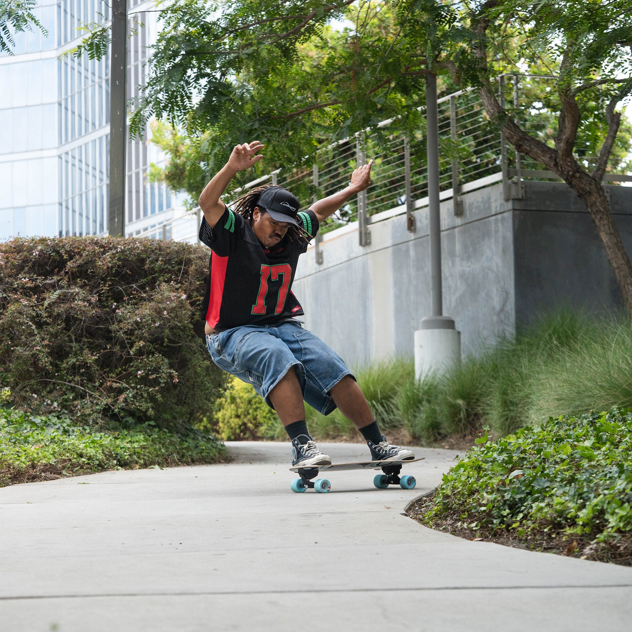 A person skateboarding outdoors on a paved path, wearing a black and red jersey with the number 17, denim shorts, and sneakers, with trees, shrubs, and a modern building in the background.