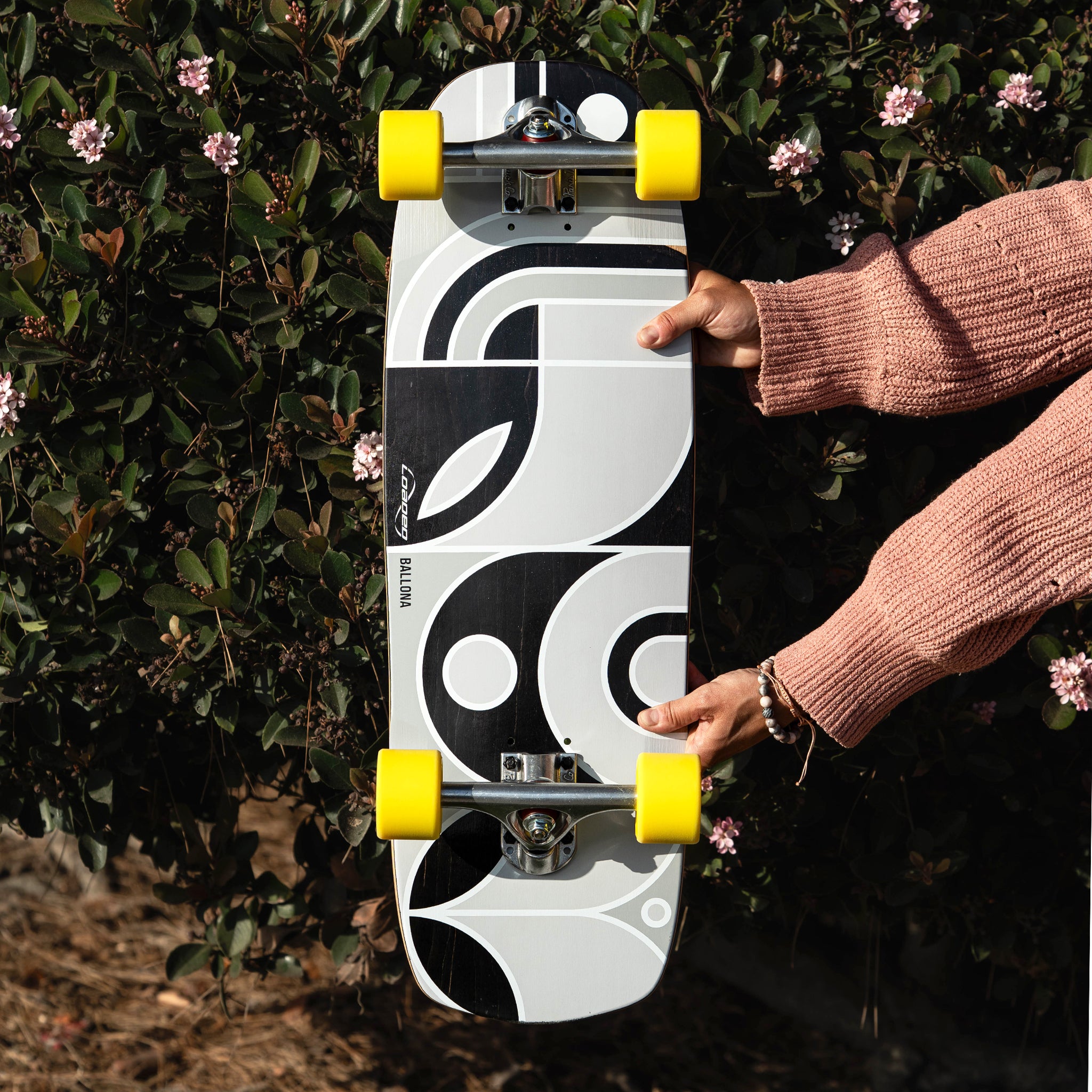 A longboard skateboard with a black, white, and gray abstract geometric pattern and bright yellow wheels, held vertically by a person wearing a pink ribbed sweater in front of a leafy green bush with small pink flowers.