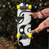 A longboard skateboard with a black, white, and gray abstract geometric pattern and bright yellow wheels, held vertically by a person wearing a pink ribbed sweater in front of a leafy green bush with small pink flowers.