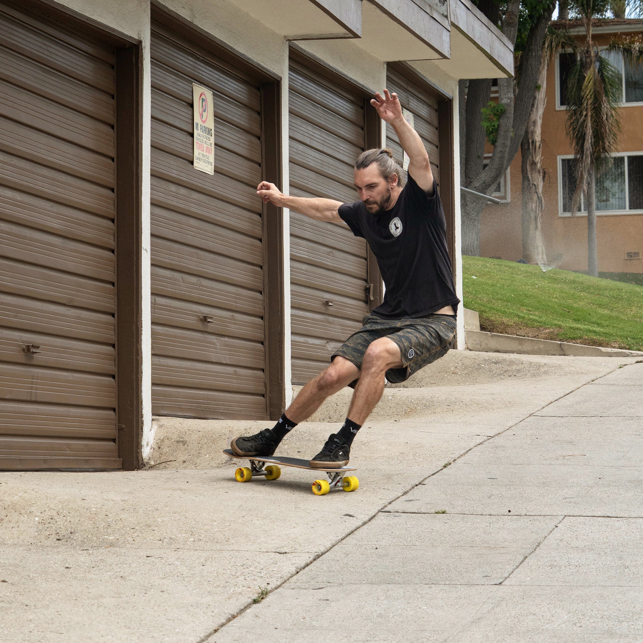 A man skateboarding on a concrete sidewalk in front of brown garage doors, wearing a black t-shirt, dark shorts, and black shoes with yellow skateboard wheels.