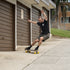 A man skateboarding on a concrete sidewalk in front of brown garage doors, wearing a black t-shirt, dark shorts, and black shoes with yellow skateboard wheels.