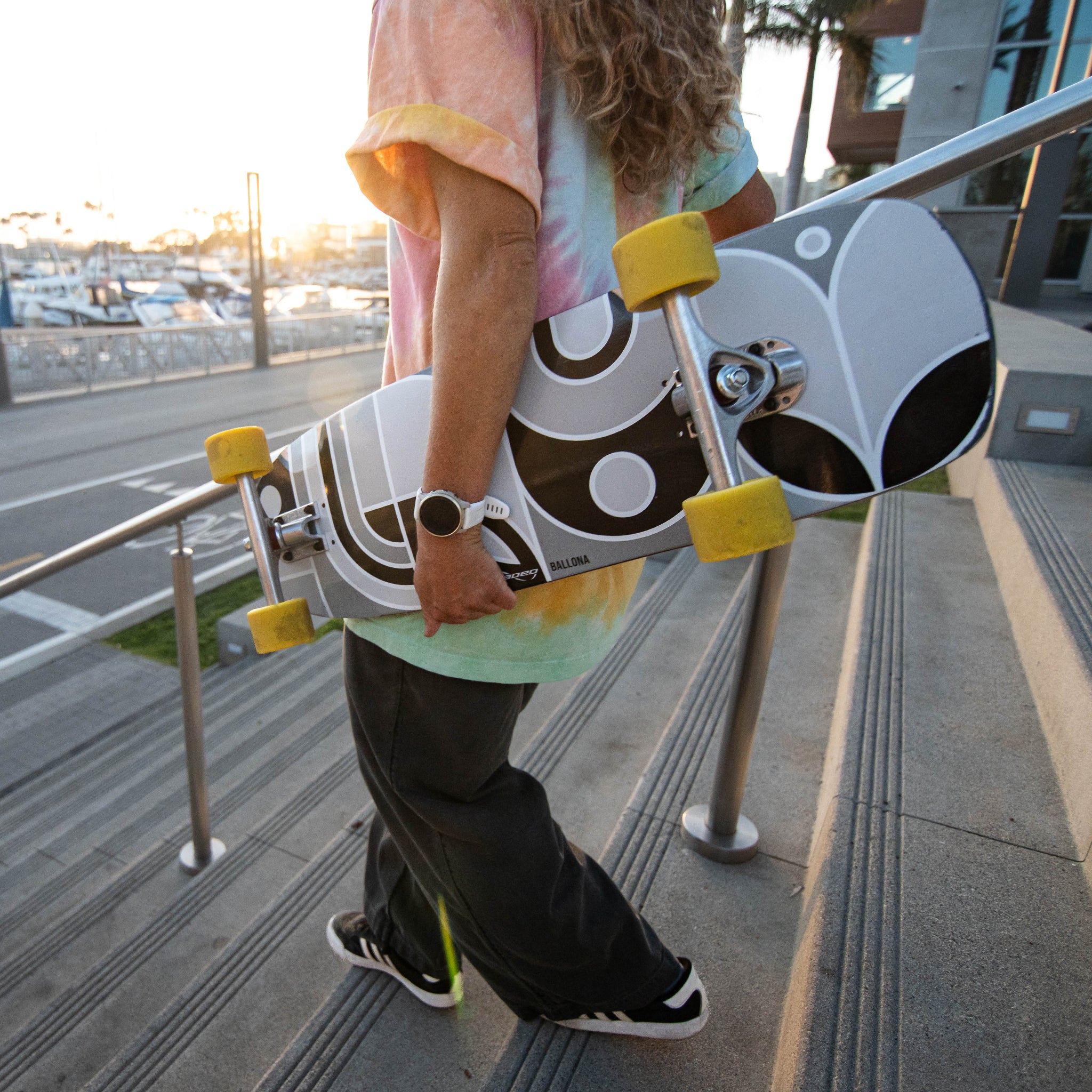 A person carrying a Loaded Ballona Orca longboard skateboard down outdoor concrete stairs at sunset. The skateboard has a white deck with a black and gray abstract graphic, yellow wheels, and silver trucks.