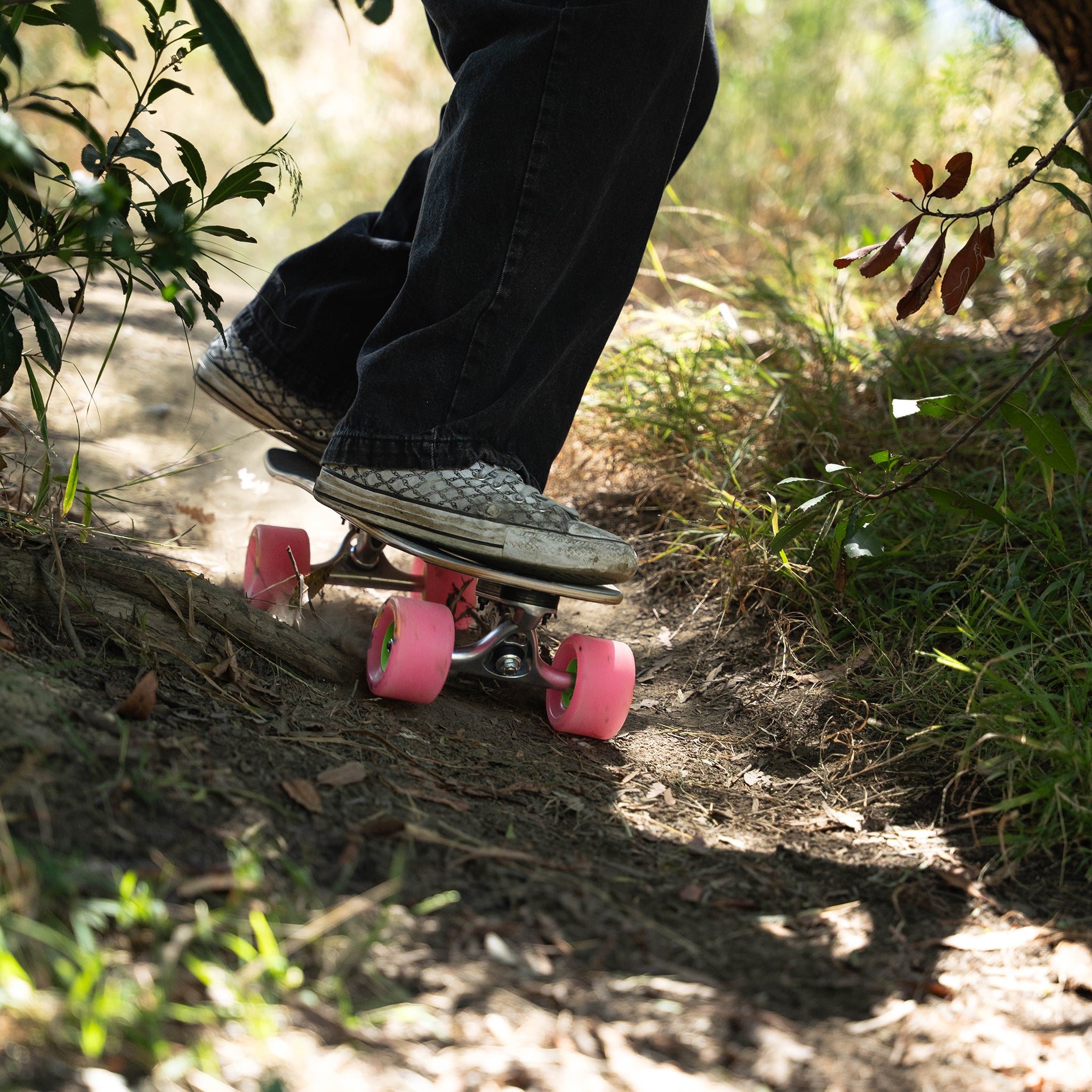 A person riding a longboard skateboard down a dirt path outdoors. The skateboard has a dark wood-grain deck, silver trucks, and bright pink wheels.
