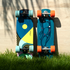 Two longboards leaning upright in grass against a sunlit stone wall. The left board has a blue, yellow, and black abstract graphic with mint wheels, and the right board has a blue abstract graphic with orange wheels.