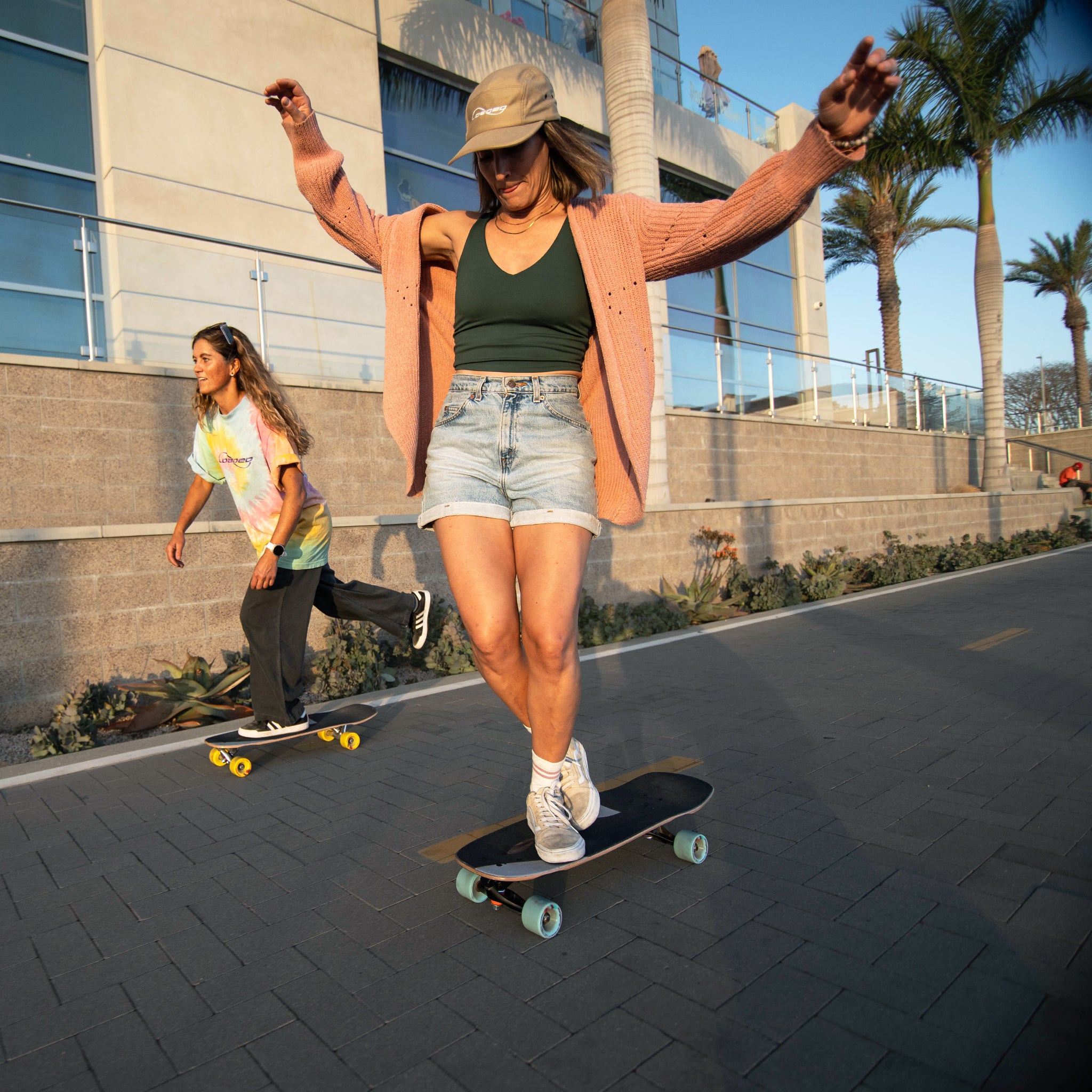 A woman skateboarding outdoors on a paved path with palm trees and a modern building in the background. She is wearing a tan bucket hat, a peach cardigan, a dark green tank top, denim shorts, and white sneakers, riding a black skateboard.