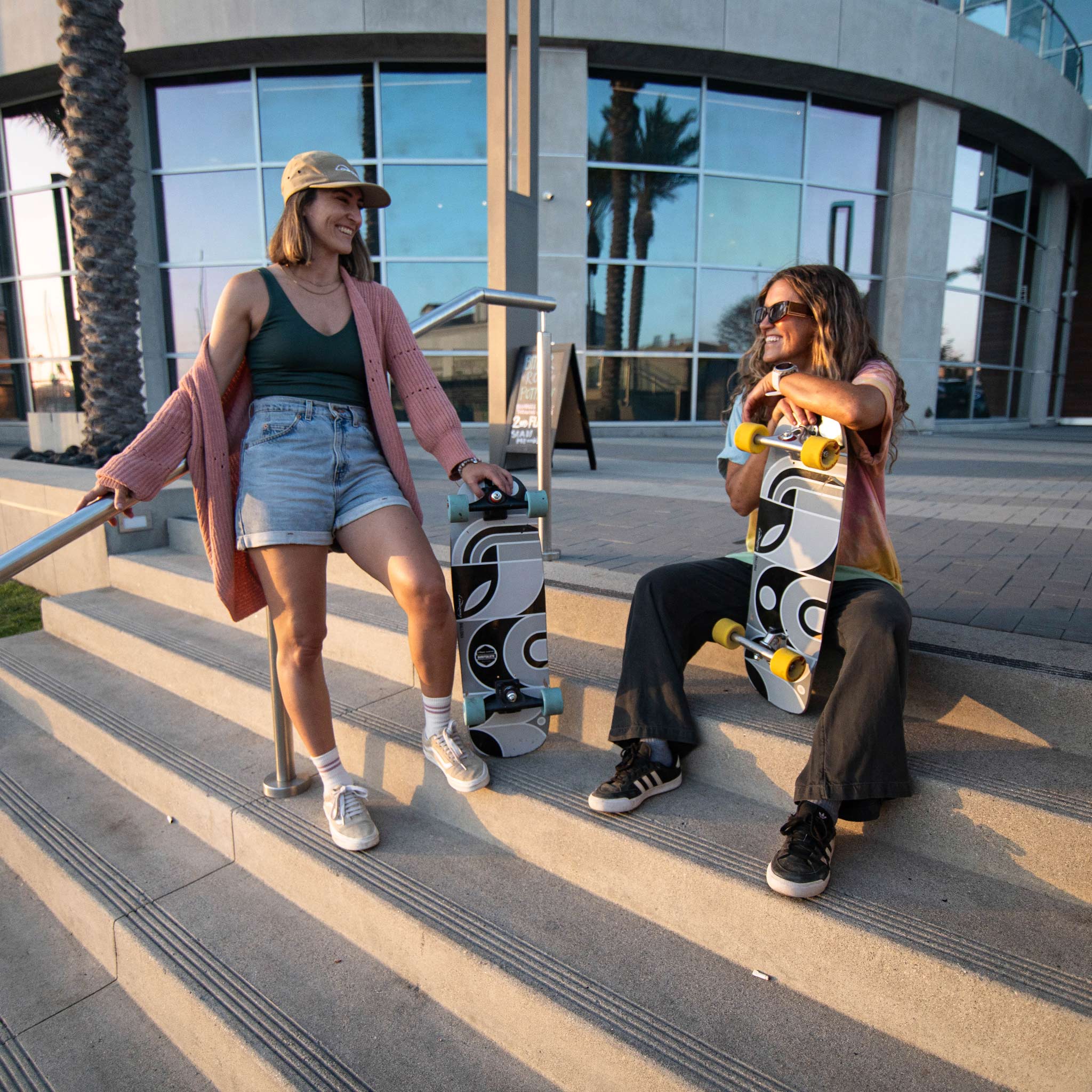 Two women on outdoor concrete steps in front of a modern glass building, each holding a skateboard with a black, white, and gray graphic design. One woman wears a pink cardigan, green tank top, denim shorts, and white sneakers, while the other wears a tie-dye shirt, black pants, and black sneakers.
