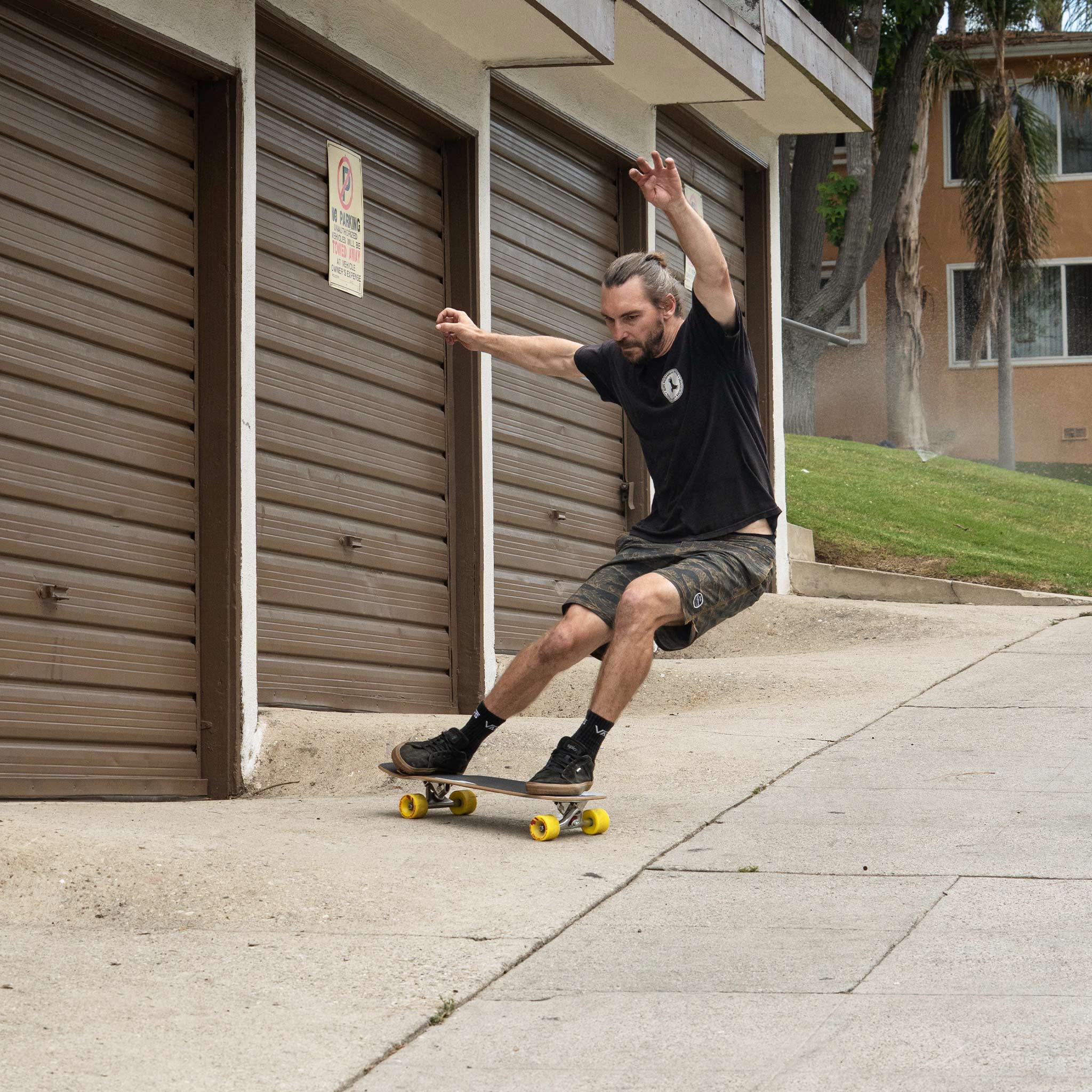 A man skateboarding on a concrete sidewalk in front of brown garage doors, wearing a black t-shirt, dark shorts, and black shoes with yellow skateboard wheels.