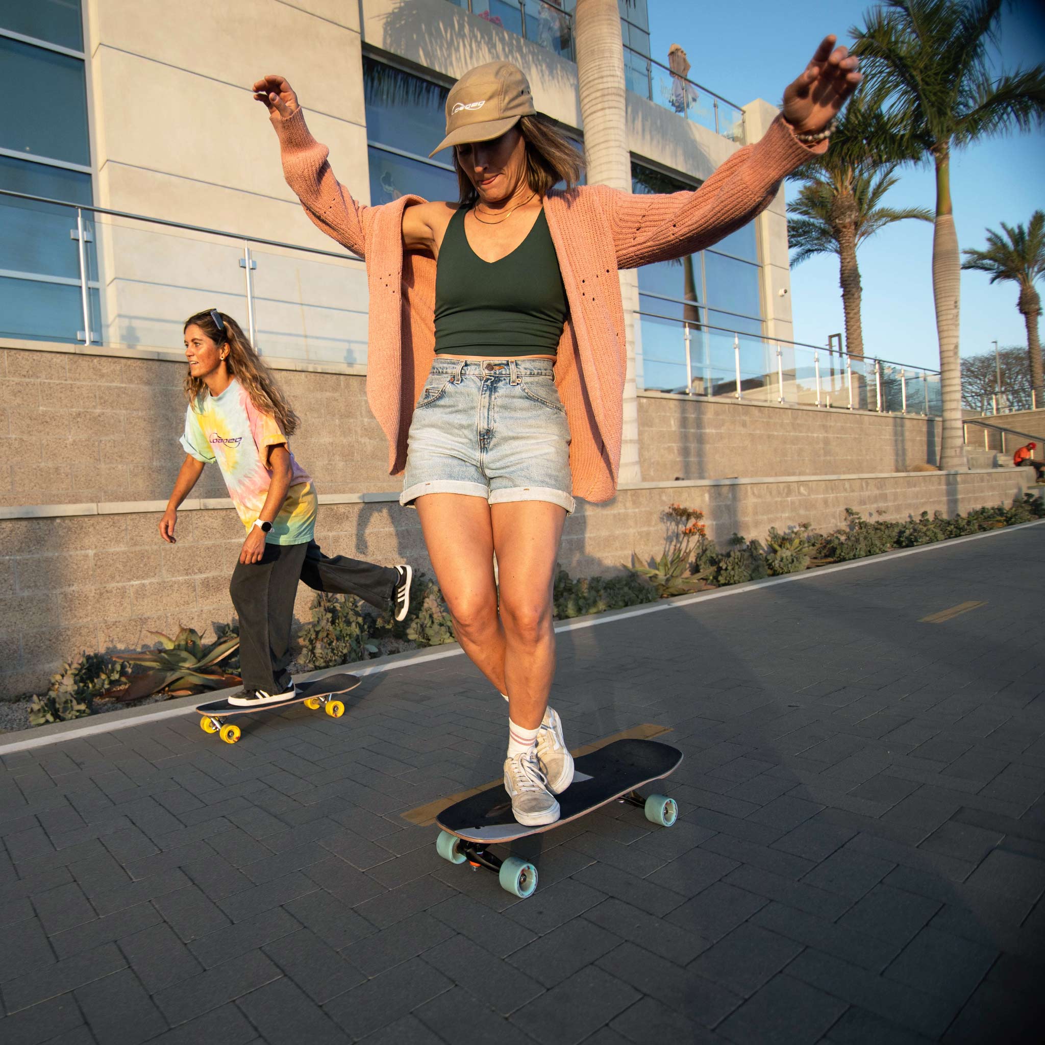A woman skateboarding outdoors on a paved path with palm trees and a modern building in the background. She is wearing a tan bucket hat, a peach cardigan, a dark green tank top, denim shorts, and white sneakers, riding a black skateboard with light blue wheels.