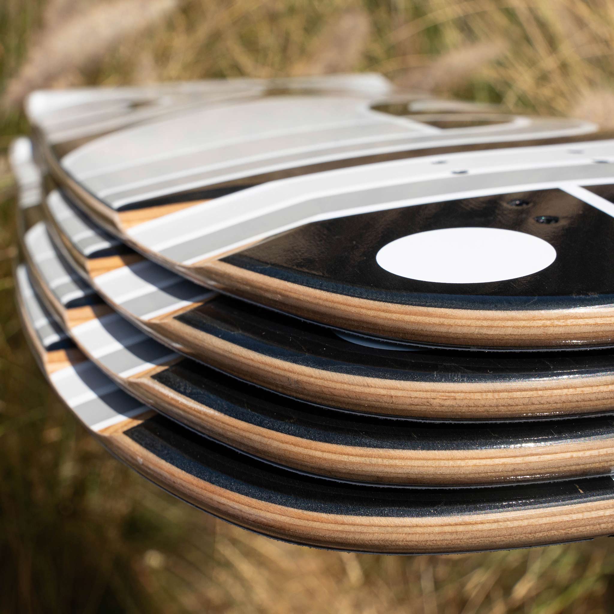 A close-up stack of skateboard decks with black grip tape, white circular stickers, and natural wood edges, photographed outdoors against a blurred grassy background.
