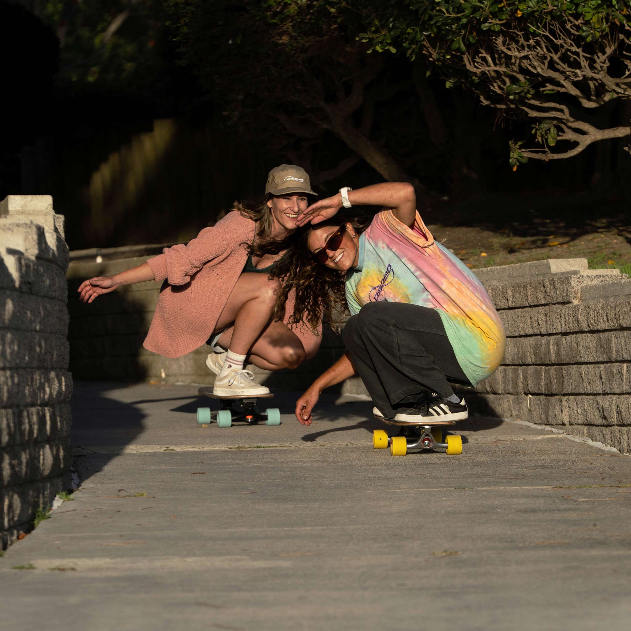 Two women crouching on longboards outdoors on a paved path with stone walls and trees in the background. One wears a pink blazer and beige cap, and the other wears a pastel rainbow tie-dye T-shirt, black pants, and white sneakers.
