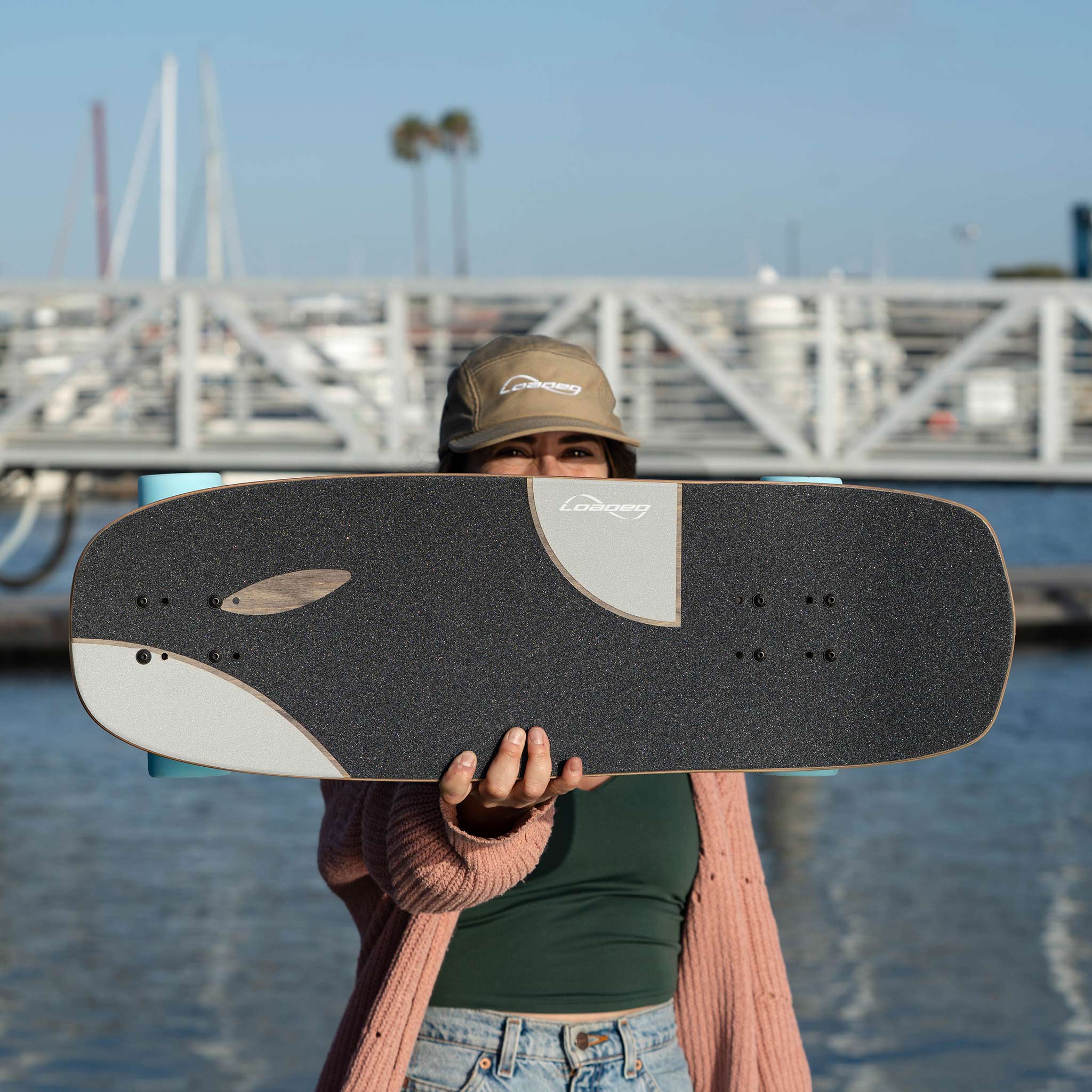 A person holding a black and white Loaded Ballona Orca skateboard in front of their face, with a marina and water in the background.