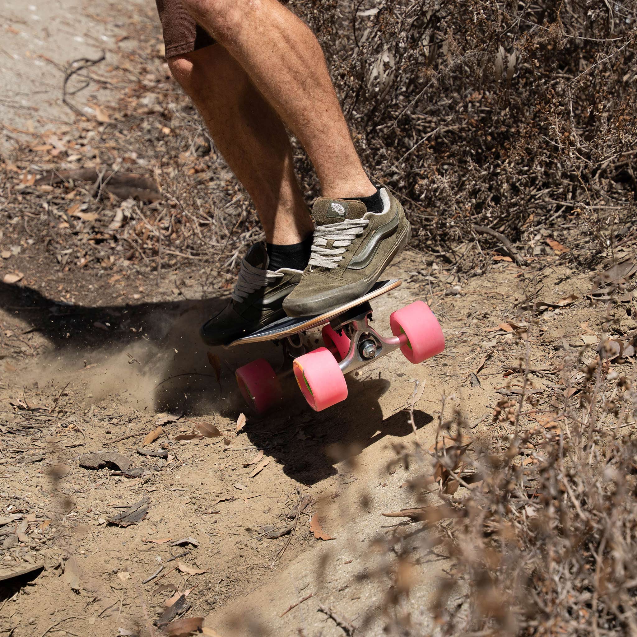 A person riding a skateboard down a dirt trail outdoors, wearing dark sneakers and shorts with bright pink wheels.