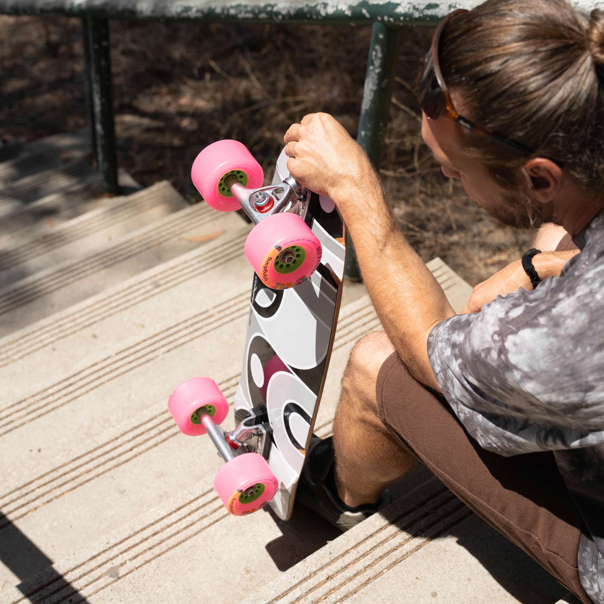 A person sitting on outdoor concrete steps holds a longboard skateboard. The skateboard has a white deck with a black and gray graphic pattern and bright pink wheels.