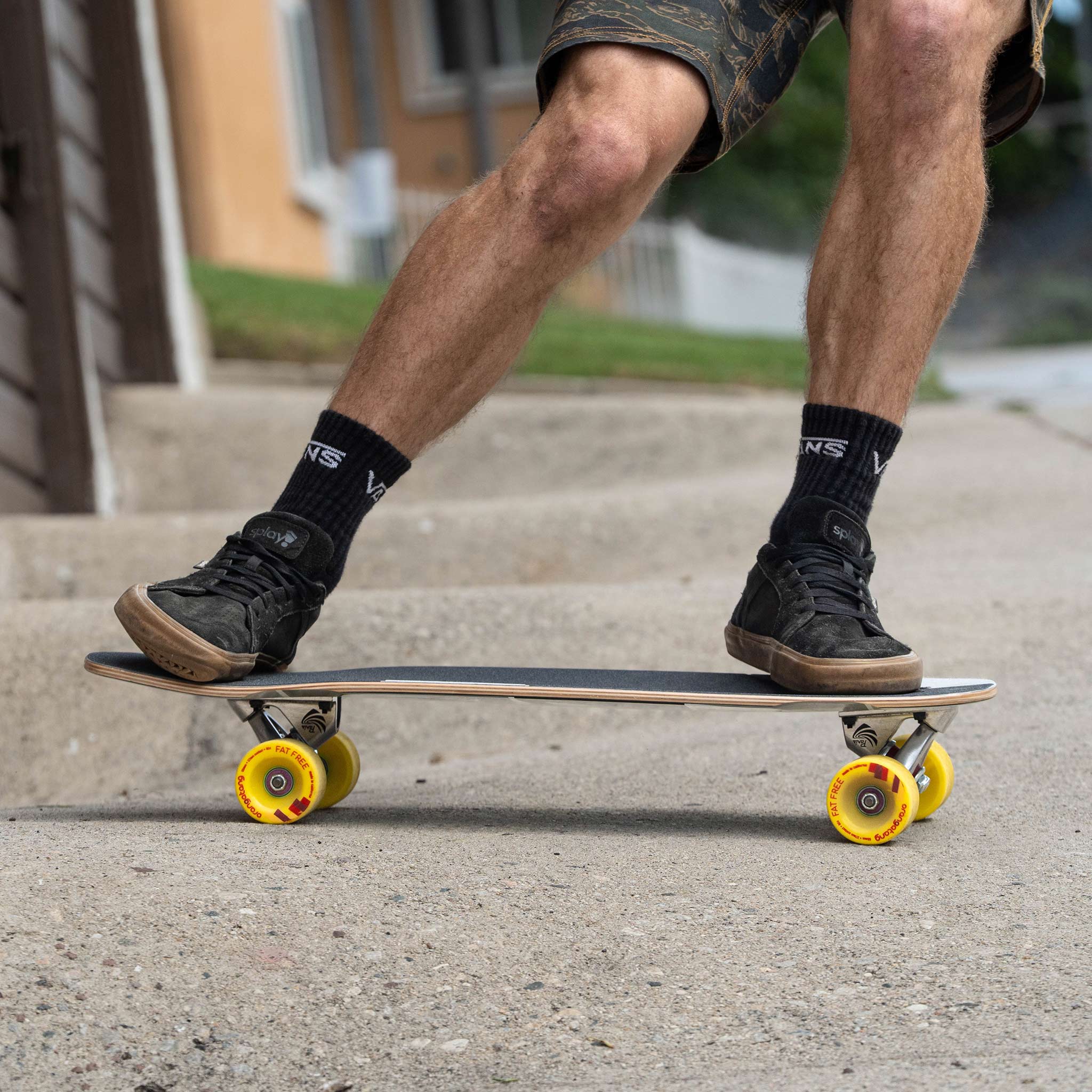 A person riding a skateboard outdoors on a concrete sidewalk, wearing black sneakers, black socks, and camouflage shorts. The skateboard has a dark wood-grain deck and bright yellow wheels.