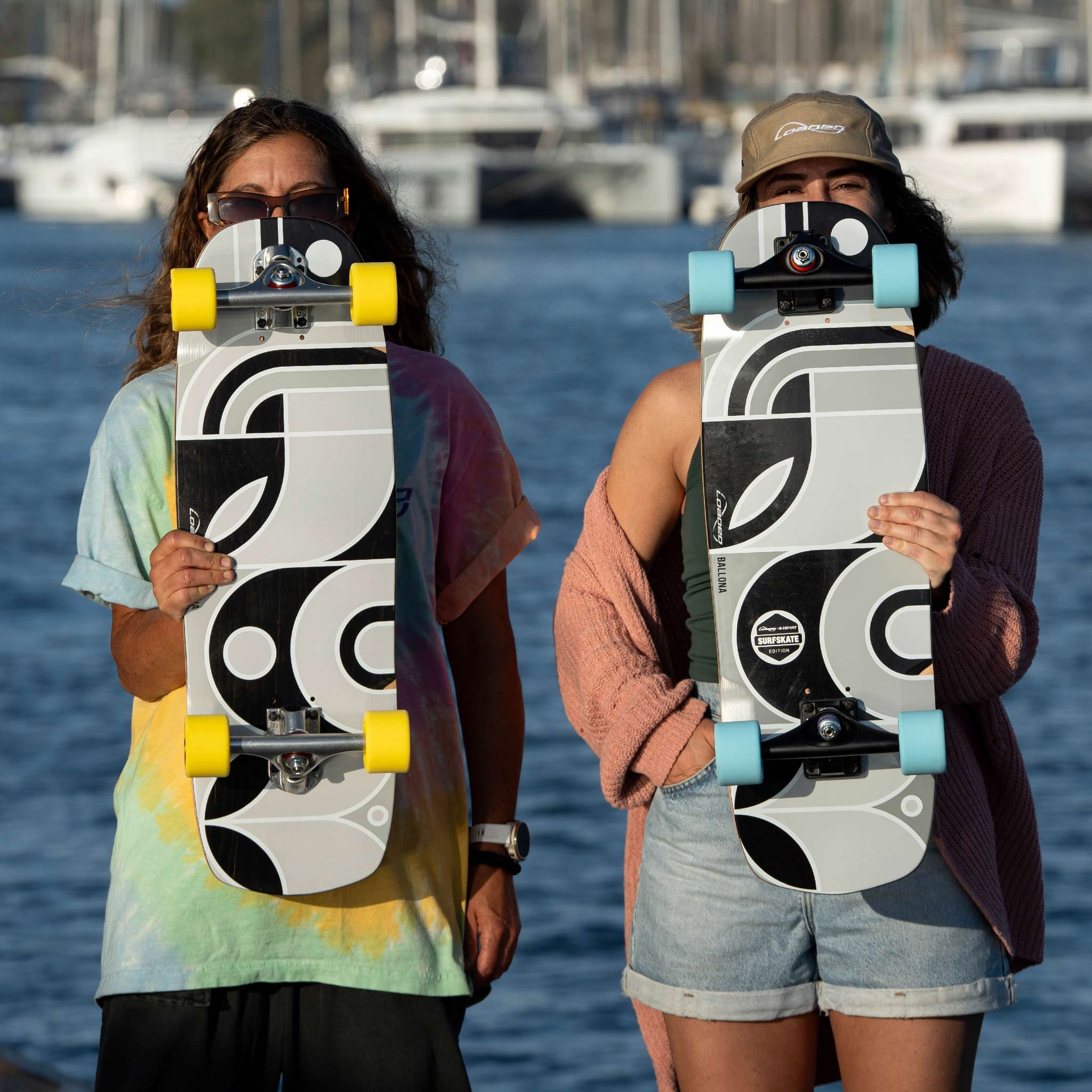 Two people standing outdoors by the water holding longboards with a matching graphic deck and contrasting yellow and light blue wheels. The background shows a blurred marina scene with boats.