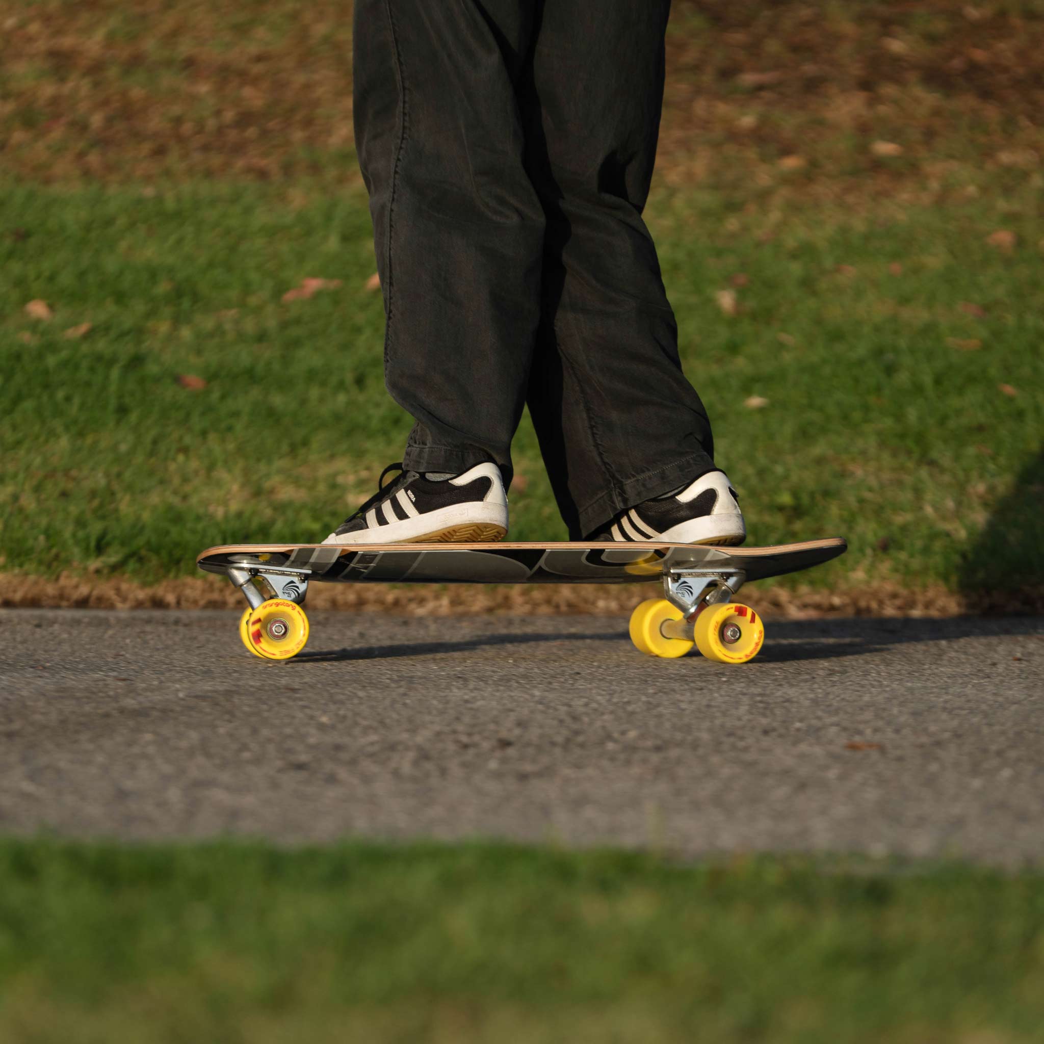 A person wearing black pants and white sneakers with black stripes stands on a skateboard with bright yellow wheels on an outdoor paved path with grass in the background.