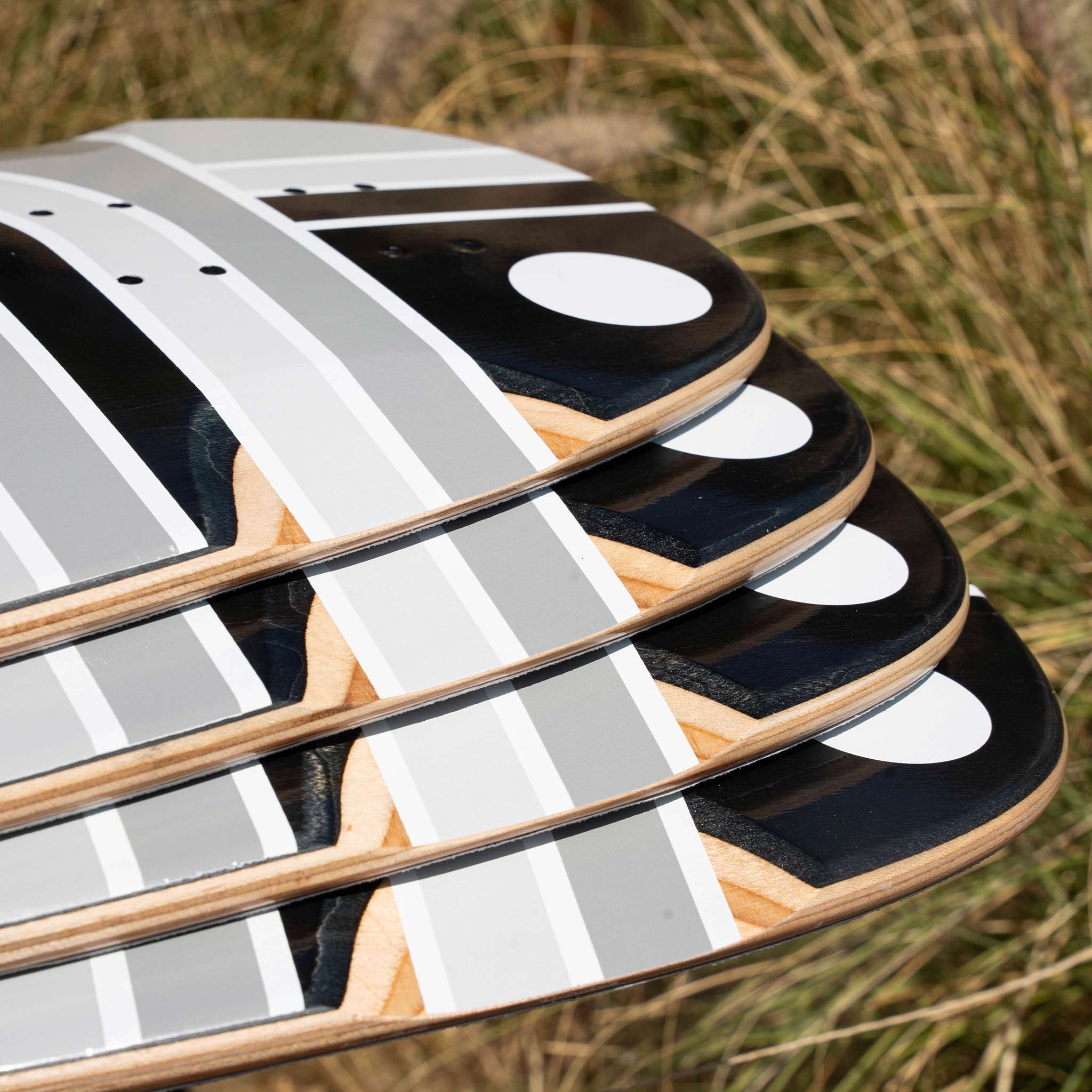 A close-up of several skateboard decks stacked in a fan arrangement, showing a black, white, and gray geometric graphic design with natural wood edges, set against a blurred background of dry grass.