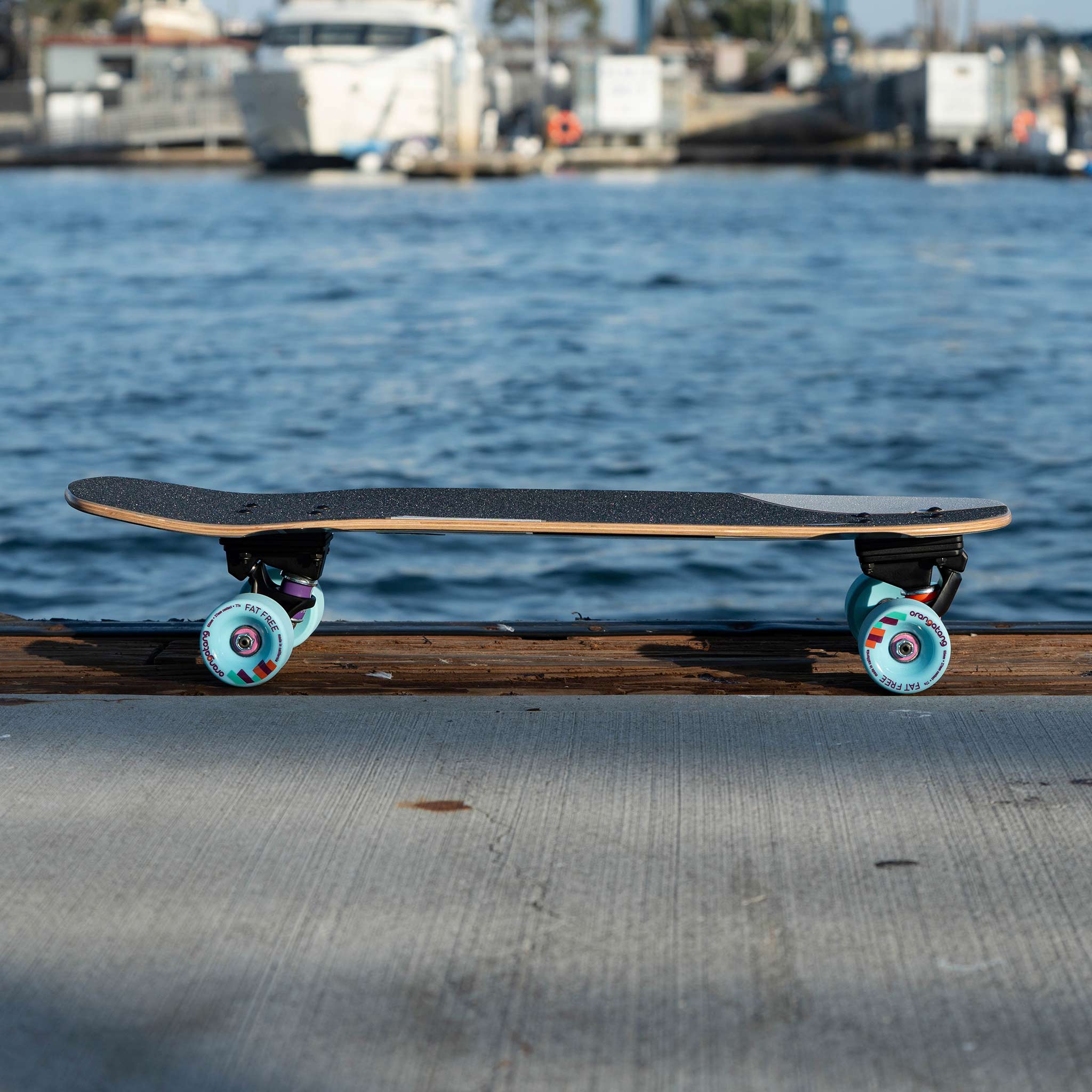 A longboard skateboard with a black grip tape deck, black trucks, and light blue wheels resting on a wooden dock with a blurred harbor and water in the background.