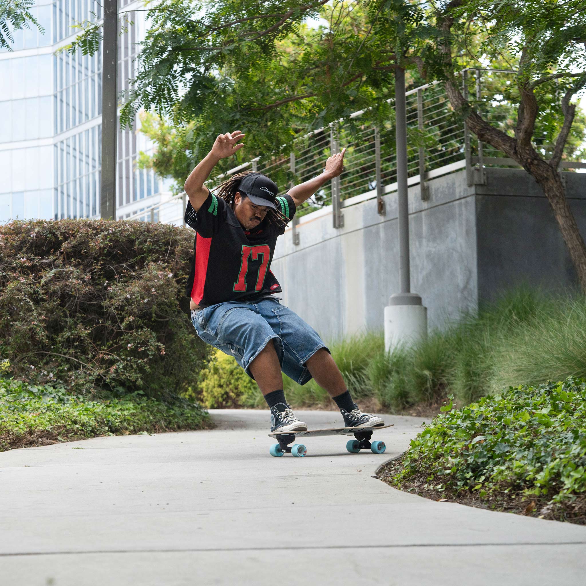 A person skateboarding outdoors on a paved path, wearing a black and red jersey with the number 17, denim shorts, and sneakers, with trees, shrubs, and a modern building in the background.
