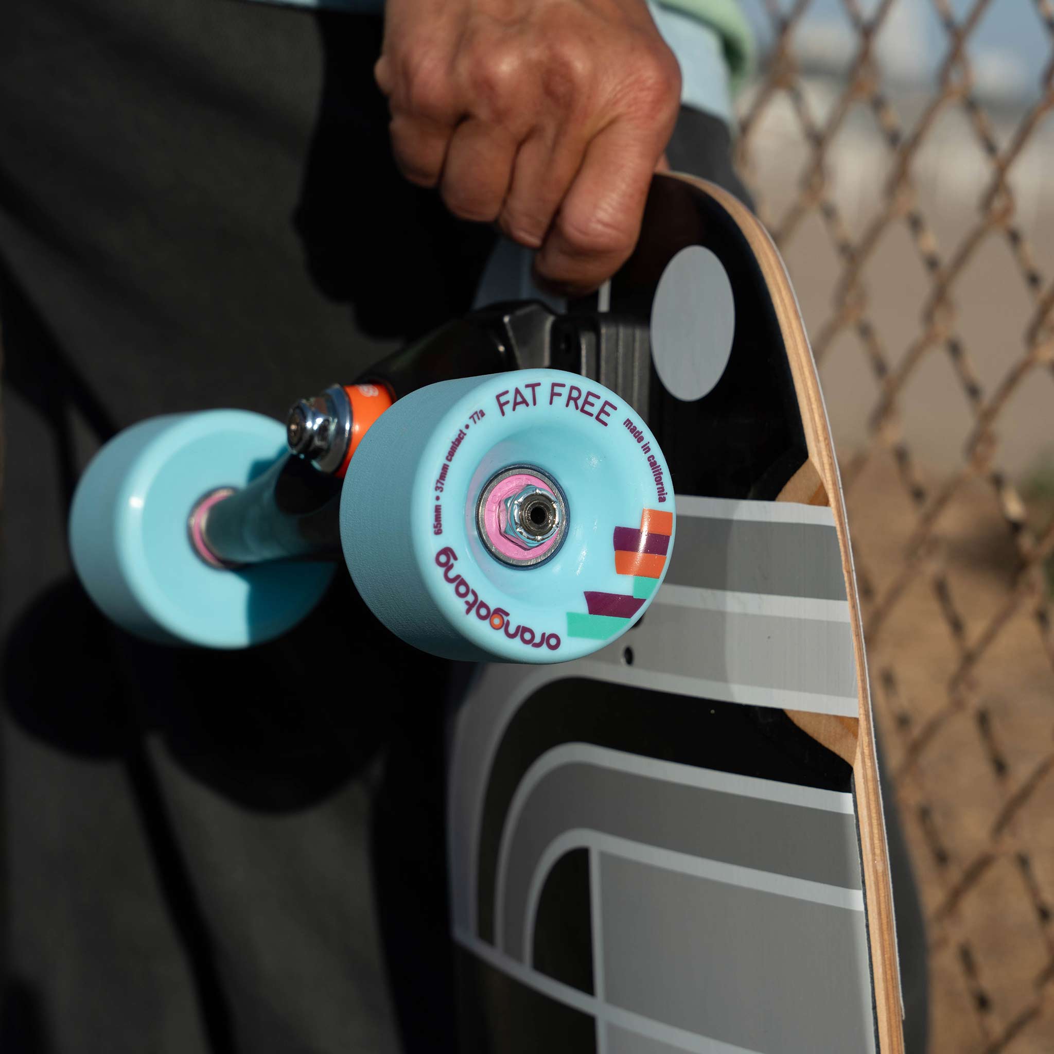 A close-up of a longboard skateboard with light blue Orangatang Fat Free wheels and a black Loaded Ballona Orca deck, held by a person outdoors near a chain-link fence. 
