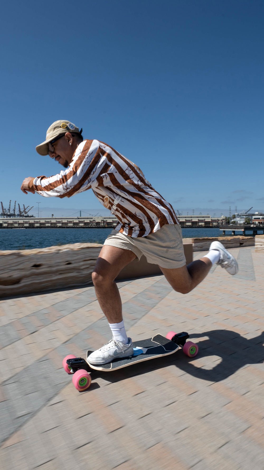 A man riding a Loaded Fathom longboard skateboard outdoors on a paved waterfront path under a clear blue sky. He is wearing a brown and white vertically striped long-sleeve shirt, beige shorts, white socks, and white sneakers with pink wheels.