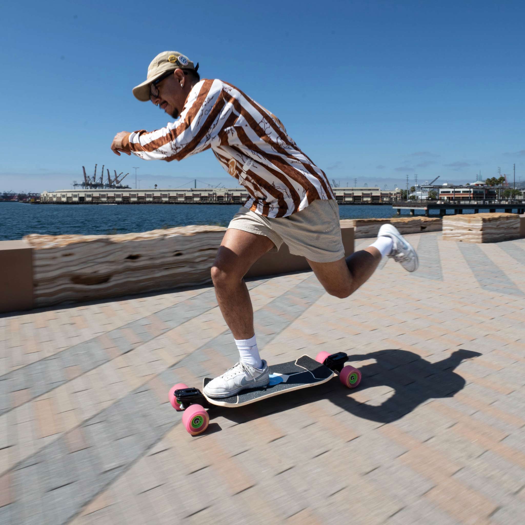 A man riding a longboard skateboard on a paved waterfront path. He is wearing a brown and white vertically striped long-sleeve shirt, beige shorts, white socks, and a tan baseball cap.