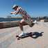 A man riding a longboard skateboard on a paved waterfront path. He is wearing a brown and white vertically striped long-sleeve shirt, beige shorts, white socks, and a tan baseball cap.