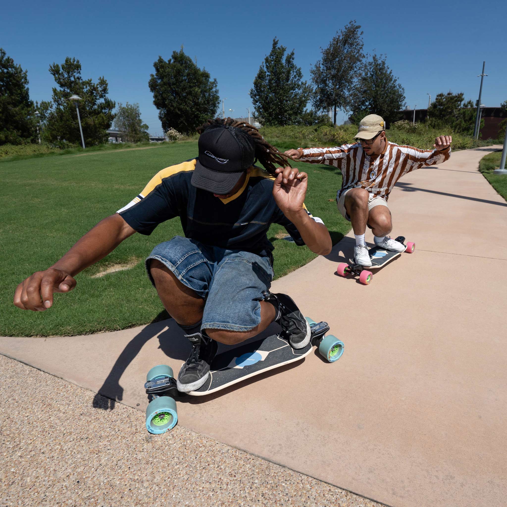 Two people skateboarding on a concrete path in a park under a clear blue sky. The person in the foreground wears a black cap, navy t-shirt, and denim shorts, while the person behind wears a brown and white striped shirt and a beige cap.