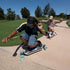 Two people skateboarding on a concrete path in a park under a clear blue sky. The person in the foreground wears a black cap, navy t-shirt, and denim shorts, while the person behind wears a brown and white striped shirt and a beige cap.