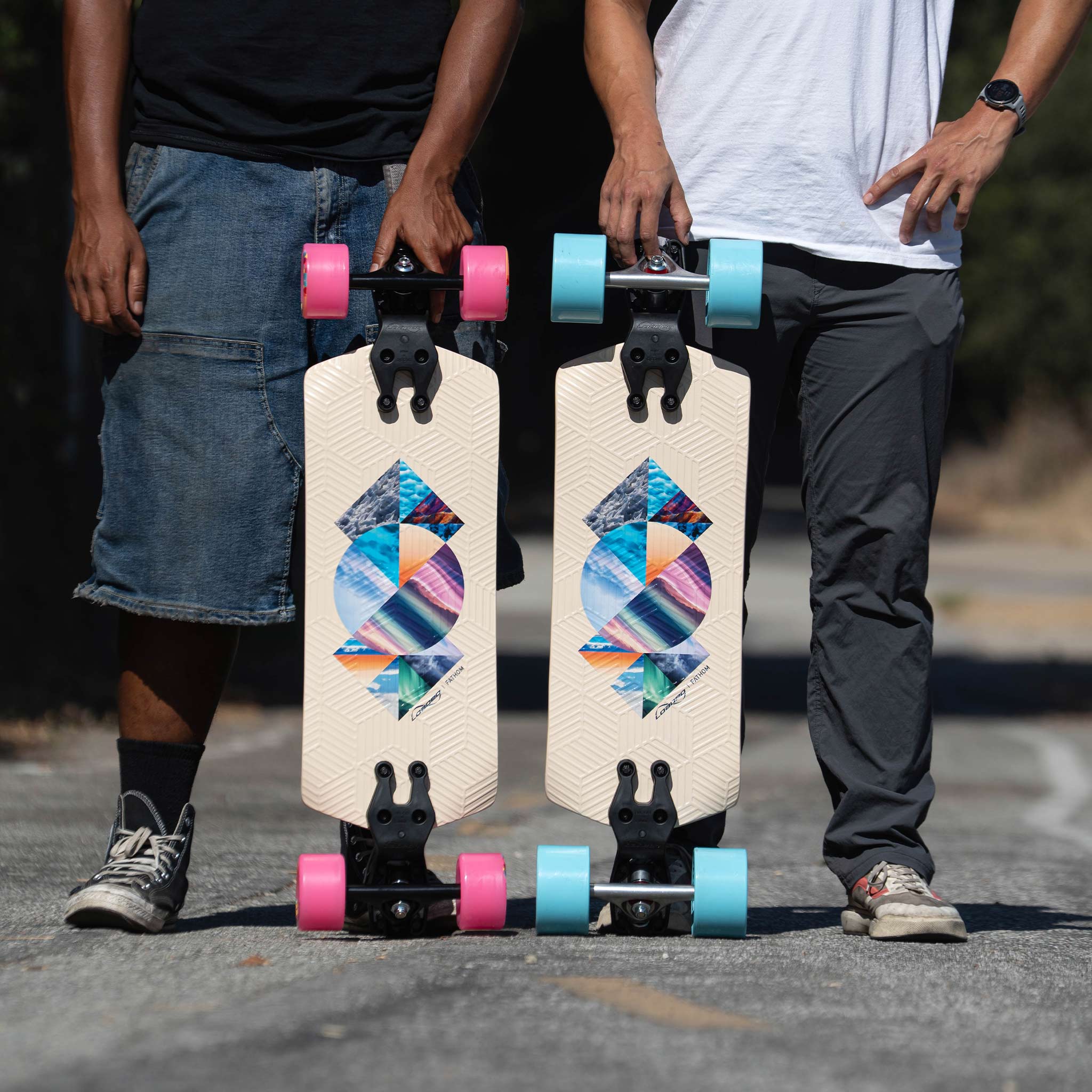 Two longboards are shown outdoors on a paved road, held upright by two partially visible people. The boards have a cream-colored deck with a colorful geometric graphic in the center, black trucks, and wheels in pink and turquoise.