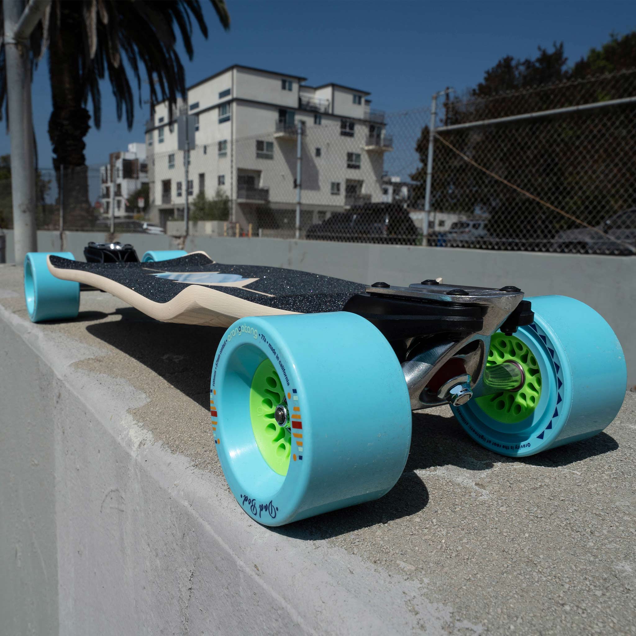 A longboard skateboard with oversized light blue wheels and bright green wheel centers, photographed outdoors on a concrete ledge with buildings and palm trees in the background.