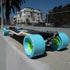A longboard skateboard with oversized light blue wheels and bright green wheel centers, photographed outdoors on a concrete ledge with buildings and palm trees in the background.