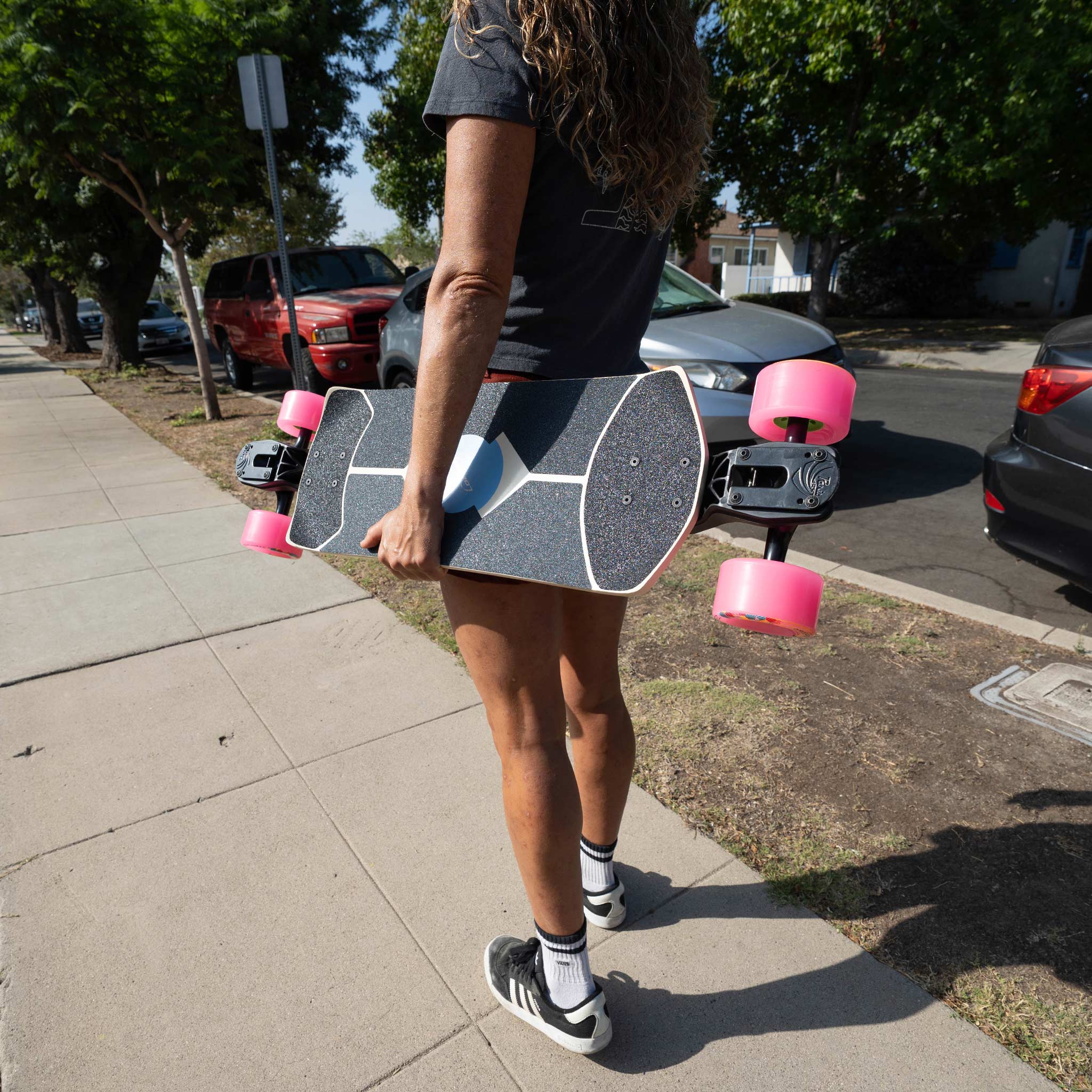 A person outdoors on a sidewalk holding a longboard skateboard. The skateboard has a black deck with white graphic lines, pink wheels, and a black handlebar with a mounted controller.