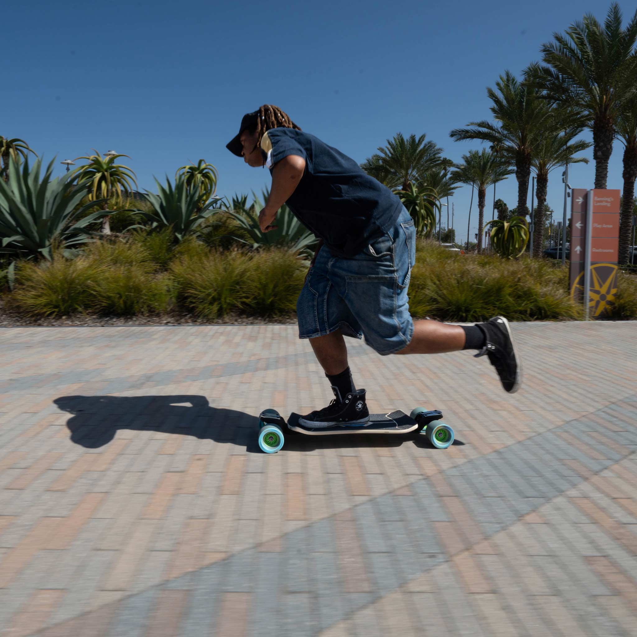 A person riding a longboard skateboard on a paved walkway outdoors, with palm trees and a blue sky in the background.