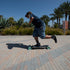 A person riding a longboard skateboard on a paved walkway outdoors, with palm trees and a blue sky in the background.