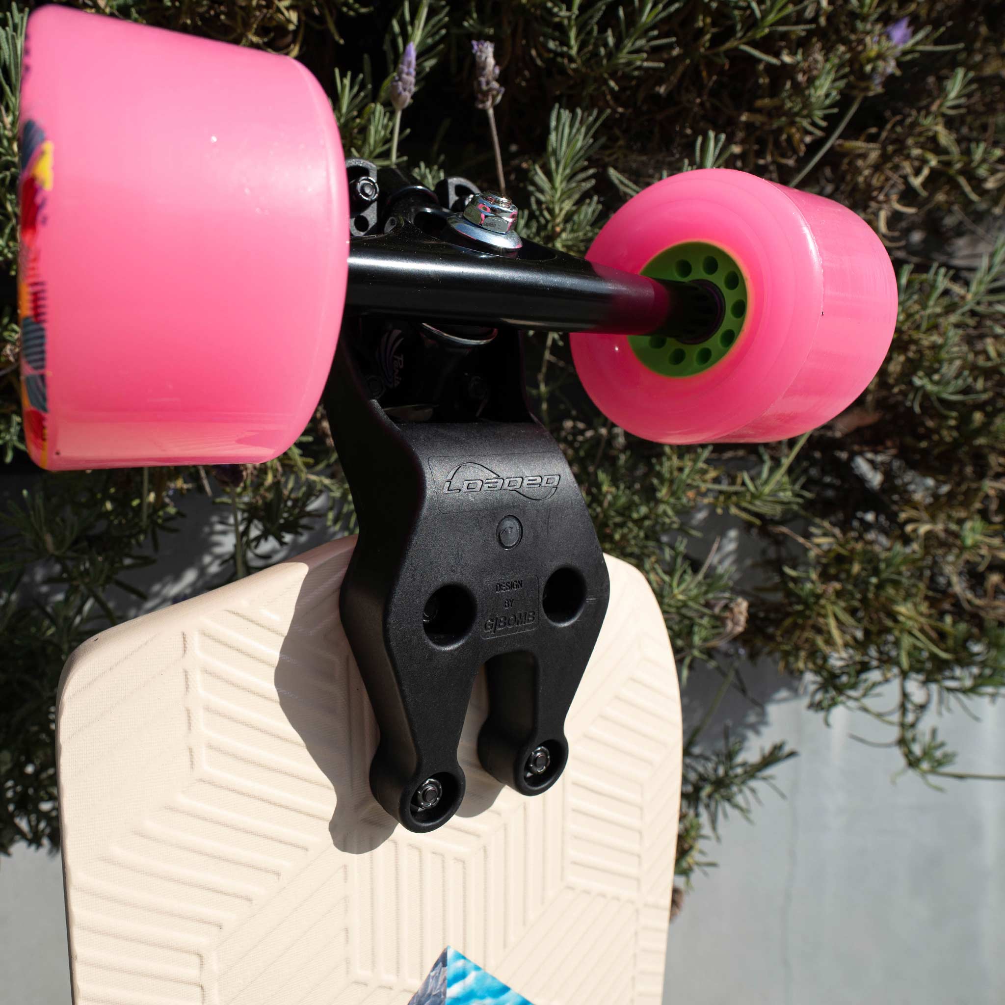 A close-up of a skateboard deck with bright pink wheels and a black truck mounted on a cream-colored deck, photographed outdoors against green foliage.