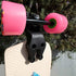 A close-up of a skateboard deck with bright pink wheels and a black truck mounted on a cream-colored deck, photographed outdoors against green foliage.
