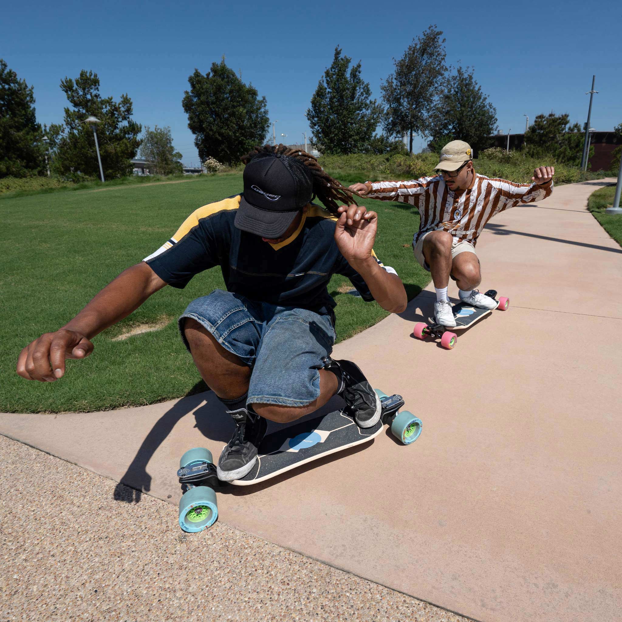 Two people skateboarding on a paved path in a park under a clear blue sky. The person in the foreground wears a black cap, dark blue t-shirt, and denim shorts, while the person behind wears a brown and white striped shirt and a beige cap.