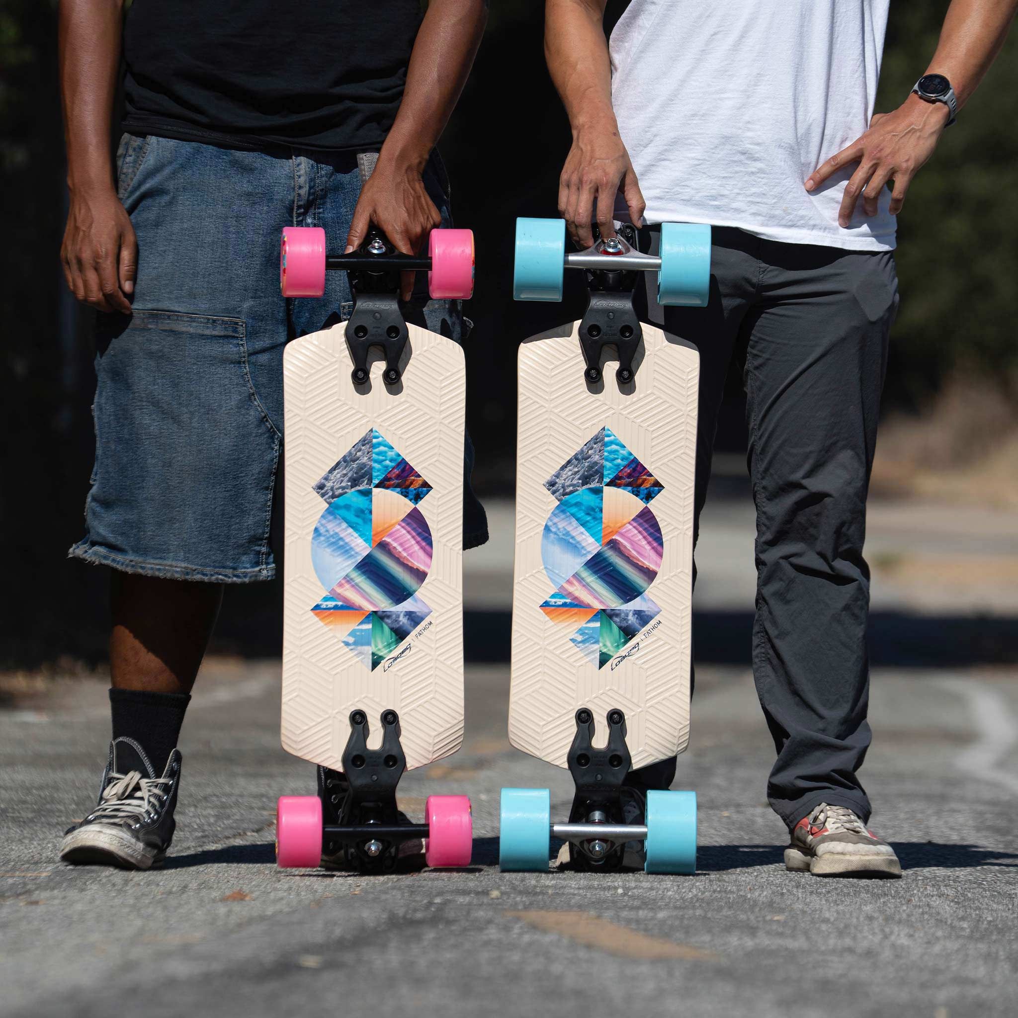 Two longboards are shown outdoors on a paved road, held upright by two partially visible people. The boards have a cream-colored deck with a colorful geometric graphic in the center, black trucks, and wheels in pink and turquoise blue.